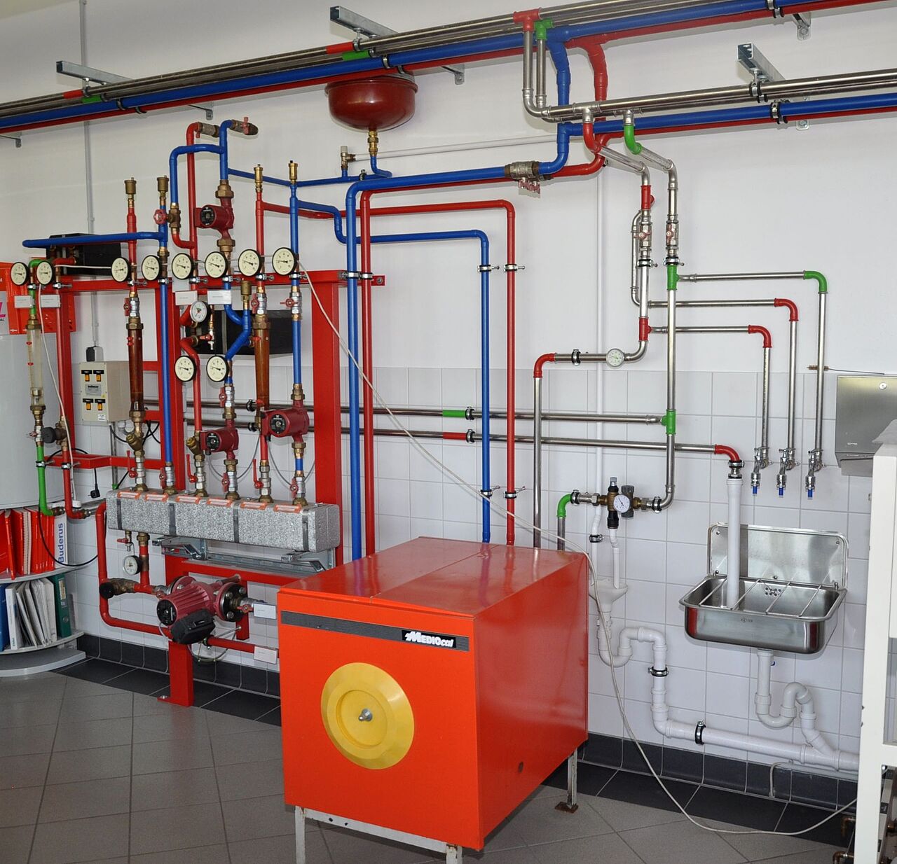Schulungsanlage mit farblich gekennzeichneten Rohrleitungen, Pumpen, Druckmessgeräten, Heizkessel und Waschbecken im SHK-Fachraum.