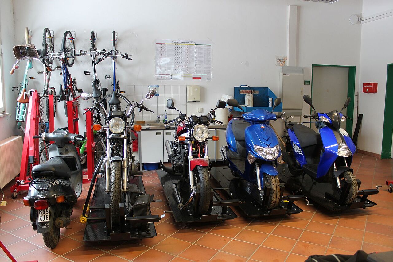 Mehrere Motorräder, Motorroller und Fahrräder stehen auf Arbeitsbühnen in der Werkstatt der Berufsfachschule Fahrzeugtechnik.