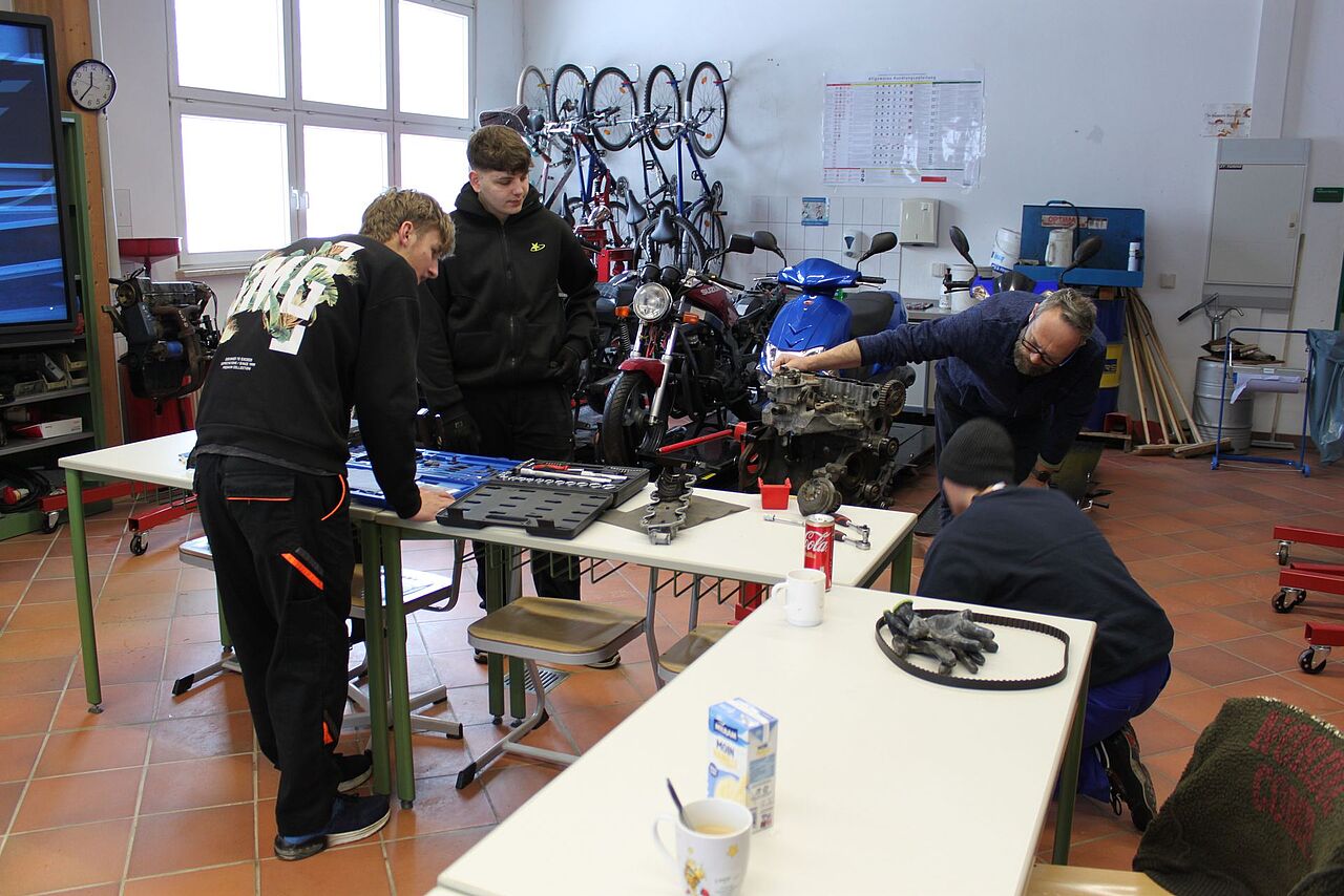 Schüler untersuchen gemeinsam mit einem Lehrer einen zerlegten Motor und verschiedene Motorteile in der Fahrzeugwerkstatt.