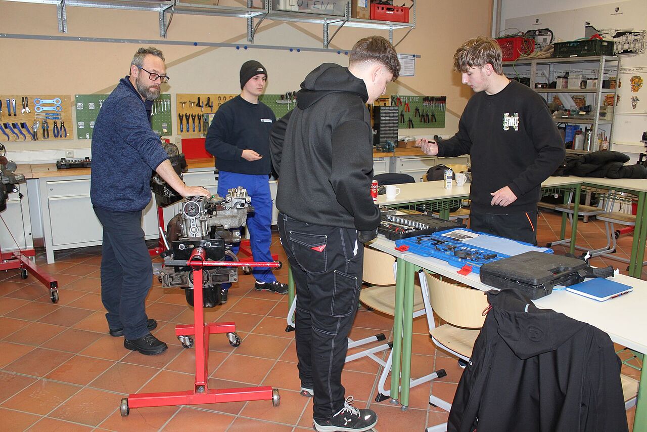 Ein Lehrer und drei Schüler arbeiten an einem ausgebauten Fahrzeugmotor und bereiten Werkzeuge für die Montage vor.