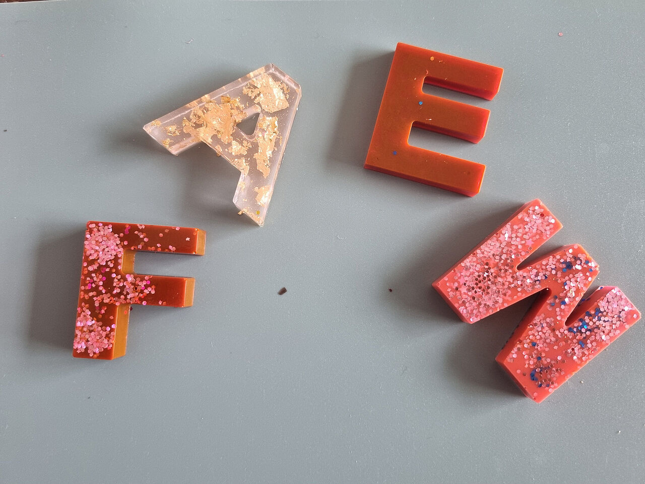 Vier dekorative Buchstaben aus rotem und transparentem Epoxidharz mit Glitzer und goldfarbenen Einschlüssen liegen auf einer Arbeitsunterlage.