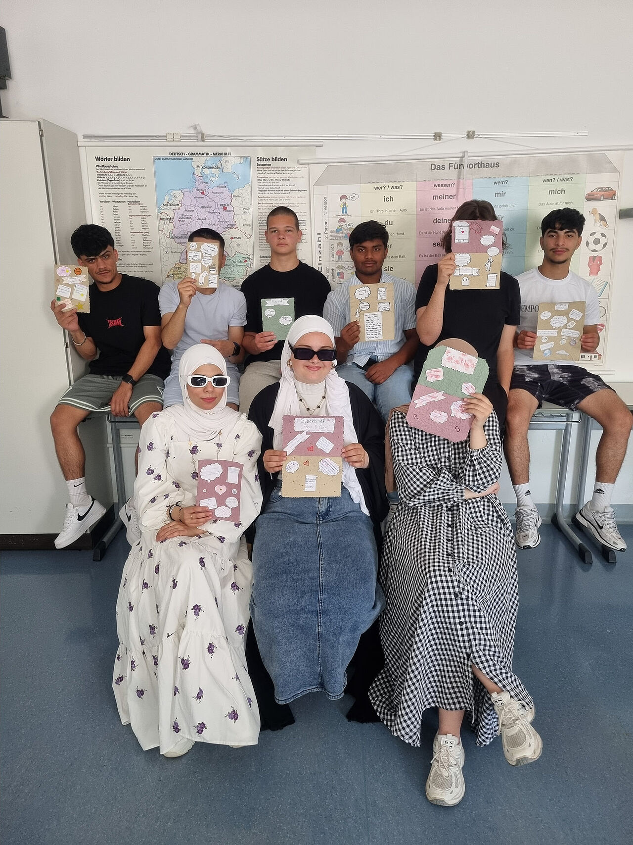 Eine Schülergruppe präsentiert selbst gestaltete Steckbriefe auf handgeschöpftem Papier vor Unterrichtstafeln zur deutschen Sprache.