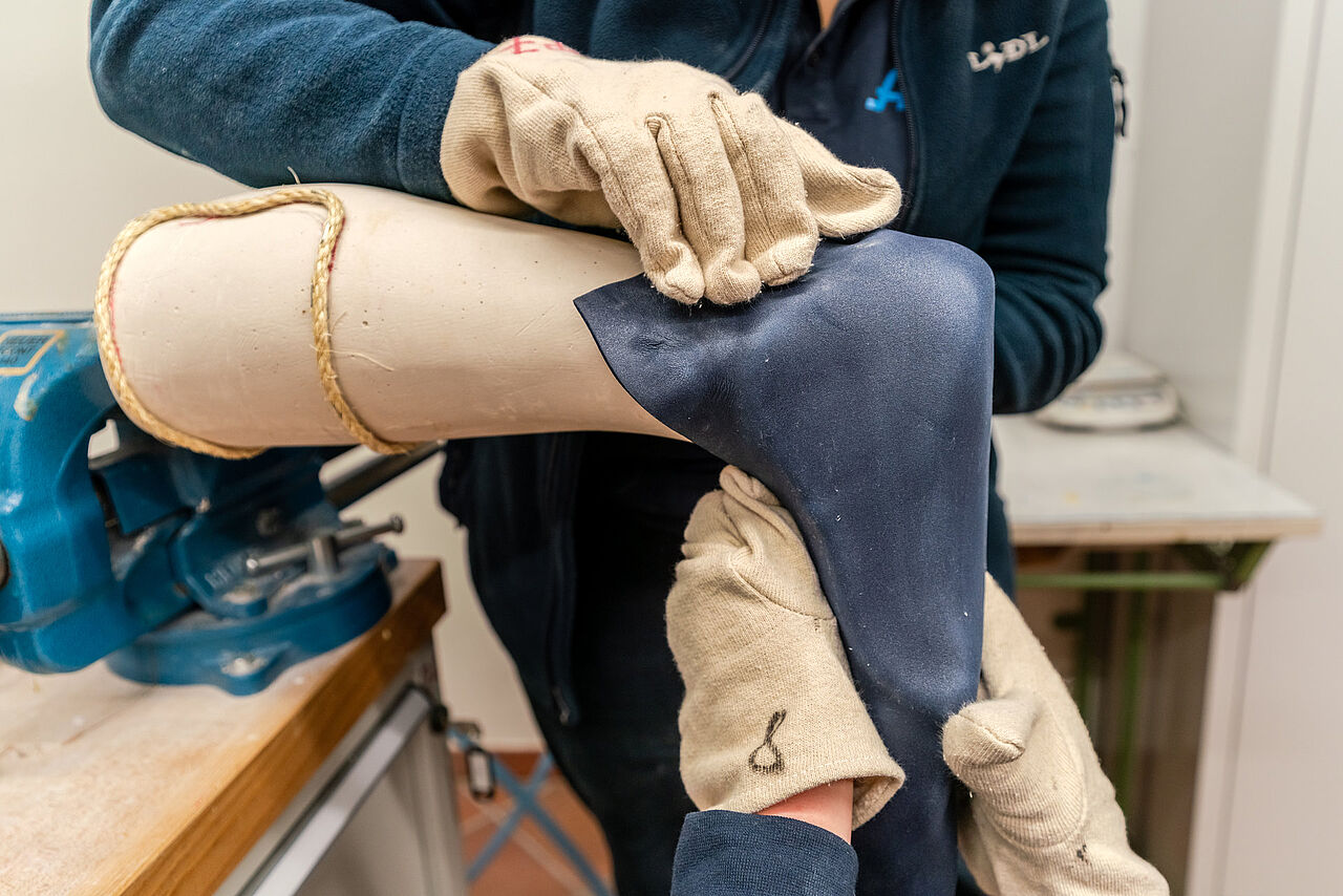 Behandschuhte Hände formen dunkles Material über einem Gipsmodell von Fuß und Unterschenkel.
