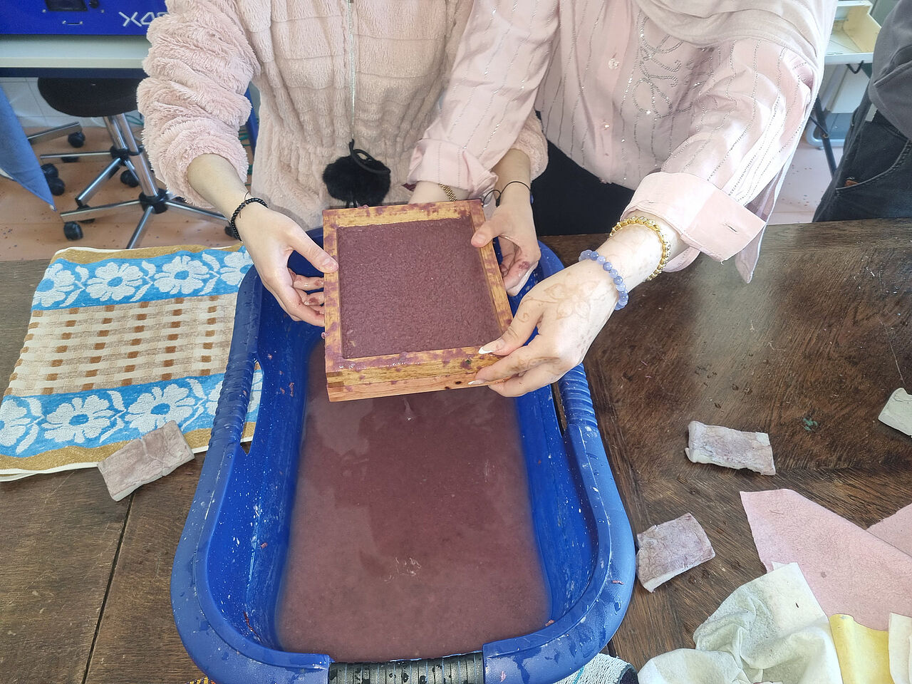 Zwei Schülerinnen heben einen Schöpfrahmen mit frisch geschöpftem violettem Papier aus einer mit Papierbrei gefüllten Wanne.