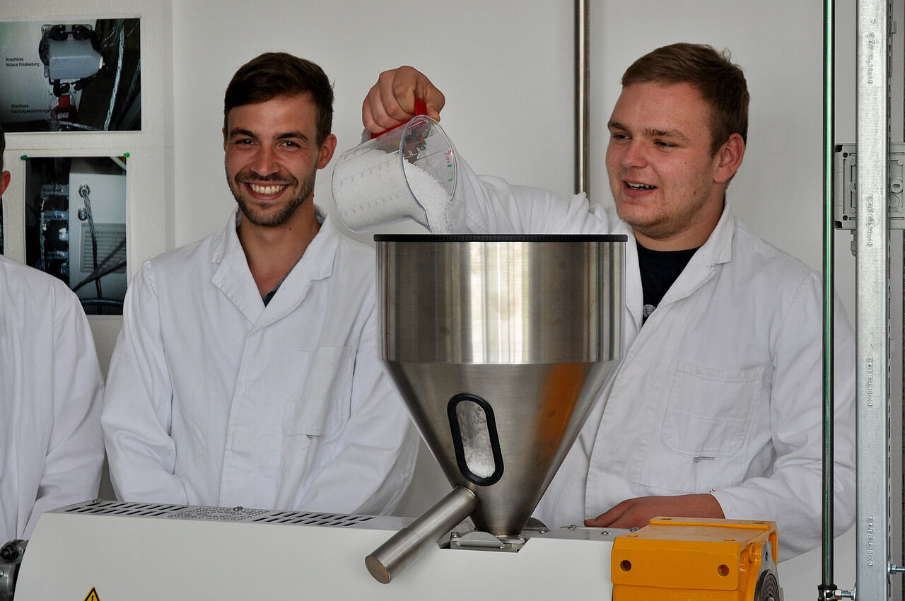 Zwei Auszubildende füllen Kunststoffgranulat in den Einfülltrichter eines Extruders.