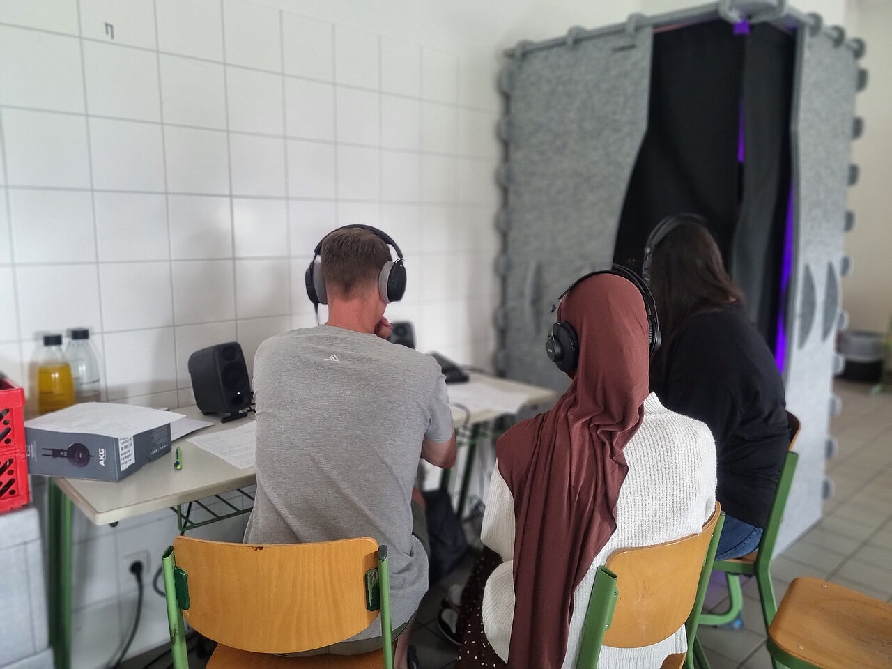Drei Projektbeteiligte hören über Kopfhörer Aufnahmen ab; im Hintergrund steht eine mobile Gesangskabine.