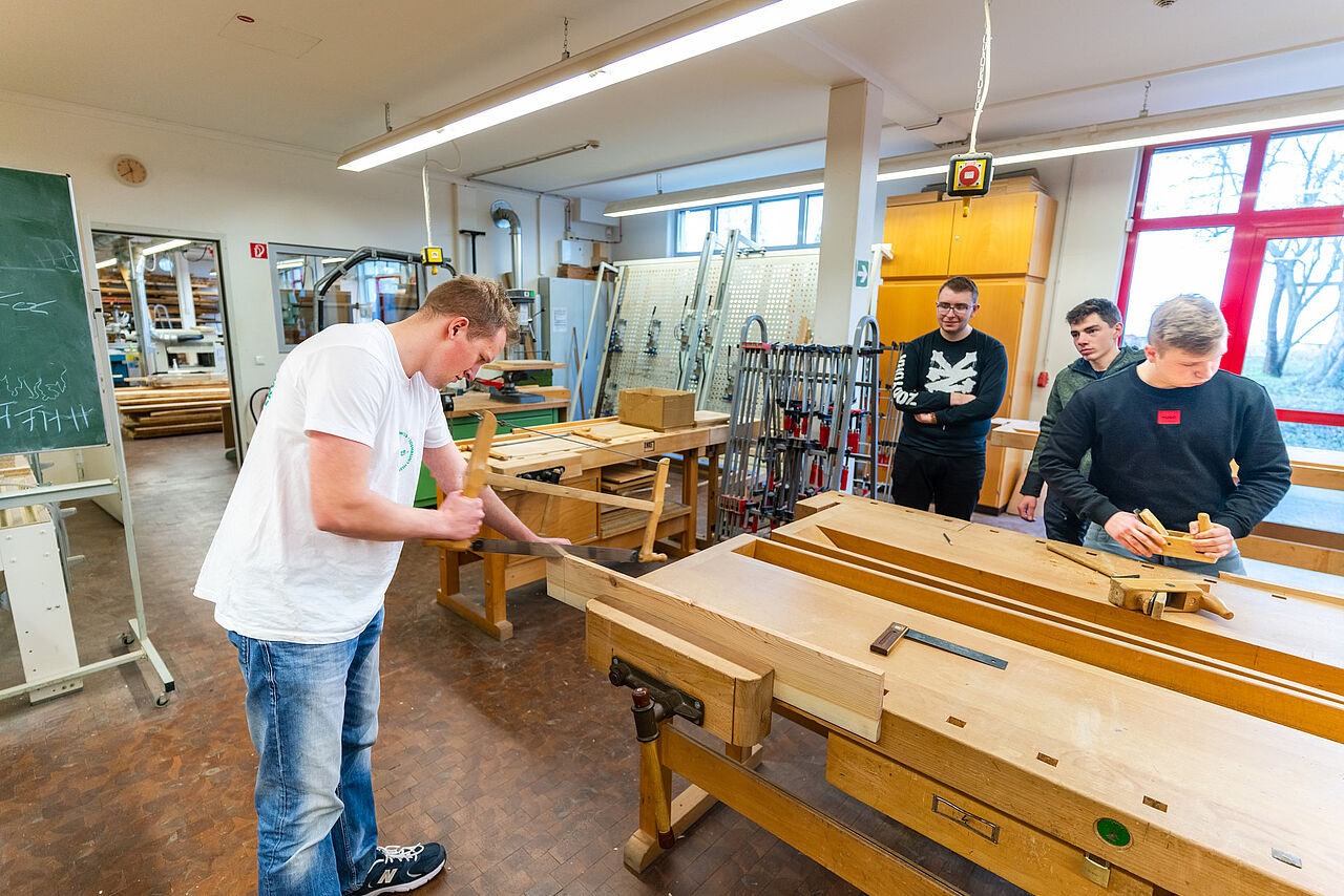 Ein Schüler demonstriert das Sägen eines Holzbretts, während Schüler die Arbeit beobachten und selbst ein Werkstück bearbeiten.