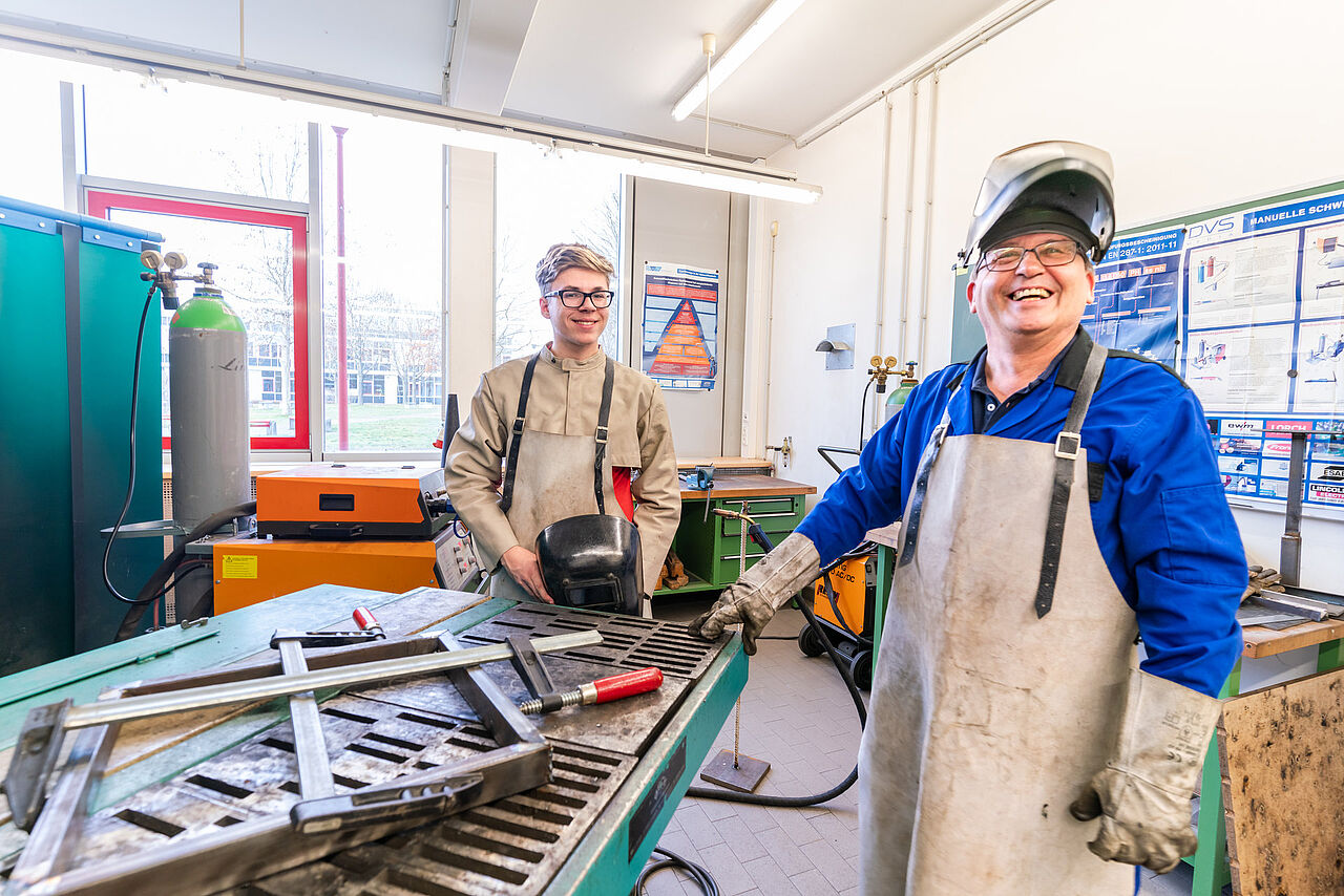Ein Auszubildender und ein Ausbilder stehen mit Schutzkleidung und Schweißhelmen an einem Arbeitstisch in der Schweißwerkstatt.