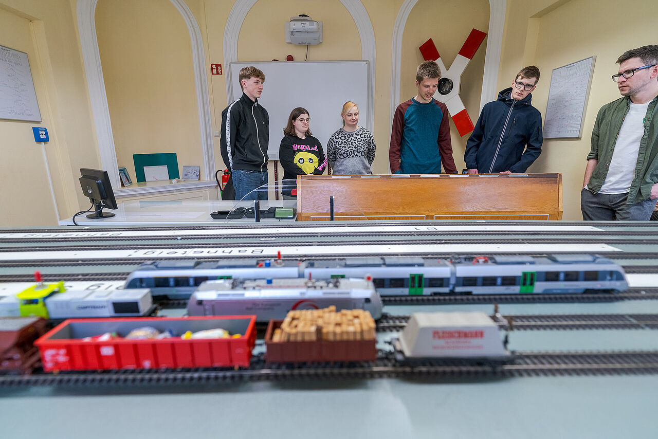 Schülerinnen und Schüler beobachten gemeinsam mit einer Lehrkraft den Betrieb auf einer Modelleisenbahnanlage.