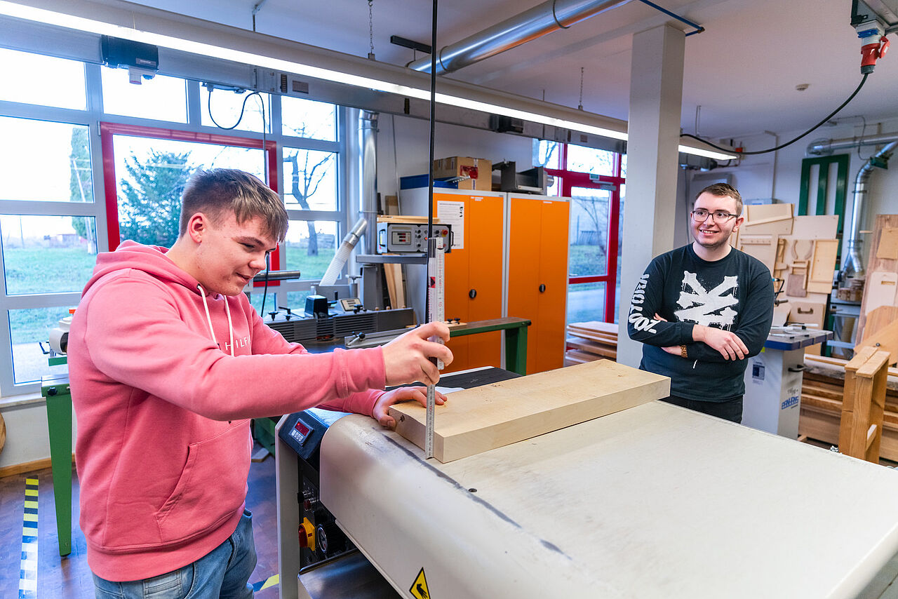 Zwei Auszubildende prüfen und vermessen ein Holzbrett auf einer Dickenhobelmaschine.