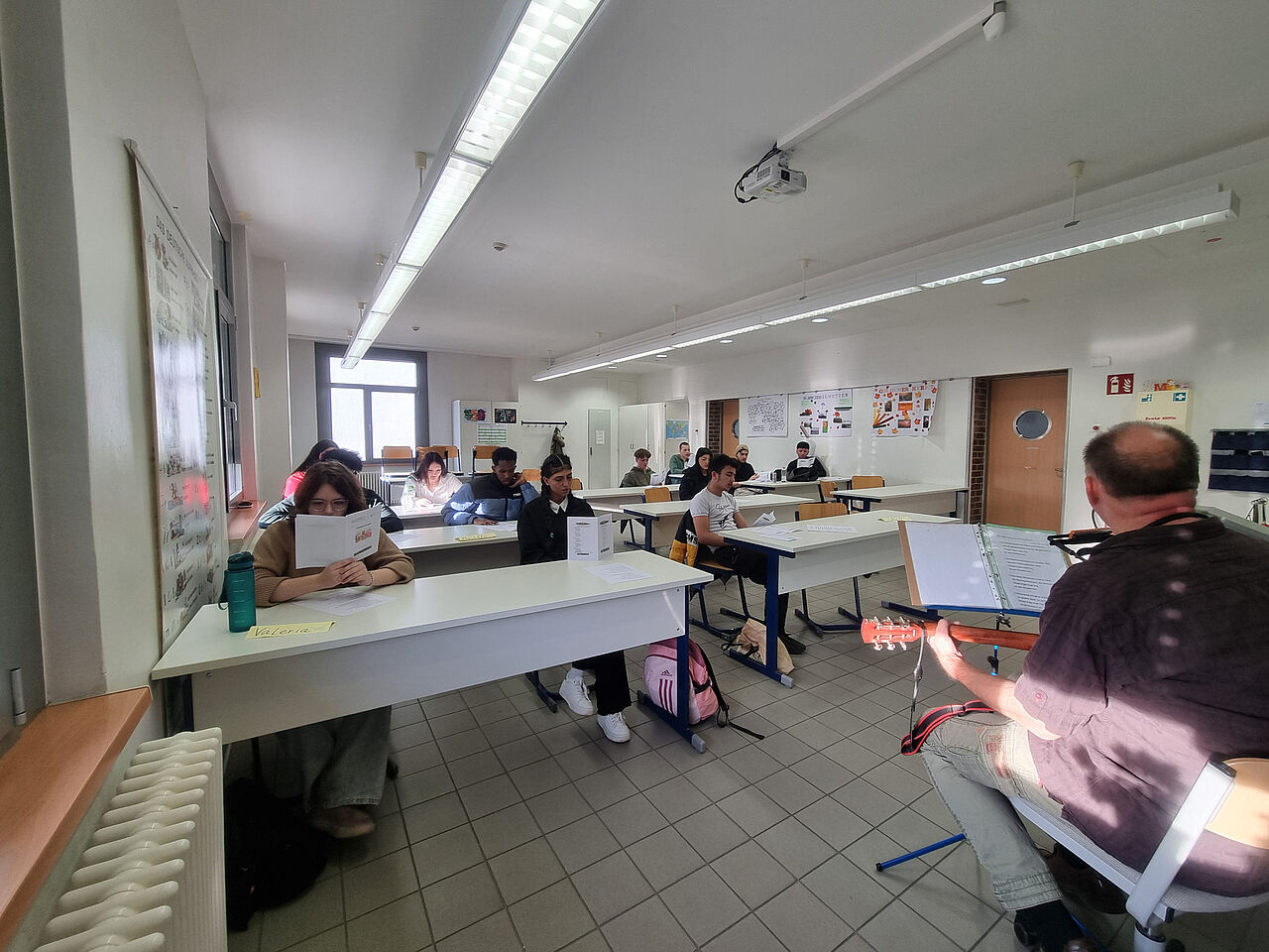 Schülerinnen und Schüler verfolgen sitzend die Liedtexte, während ein Projektleiter sie auf der Gitarre begleitet.