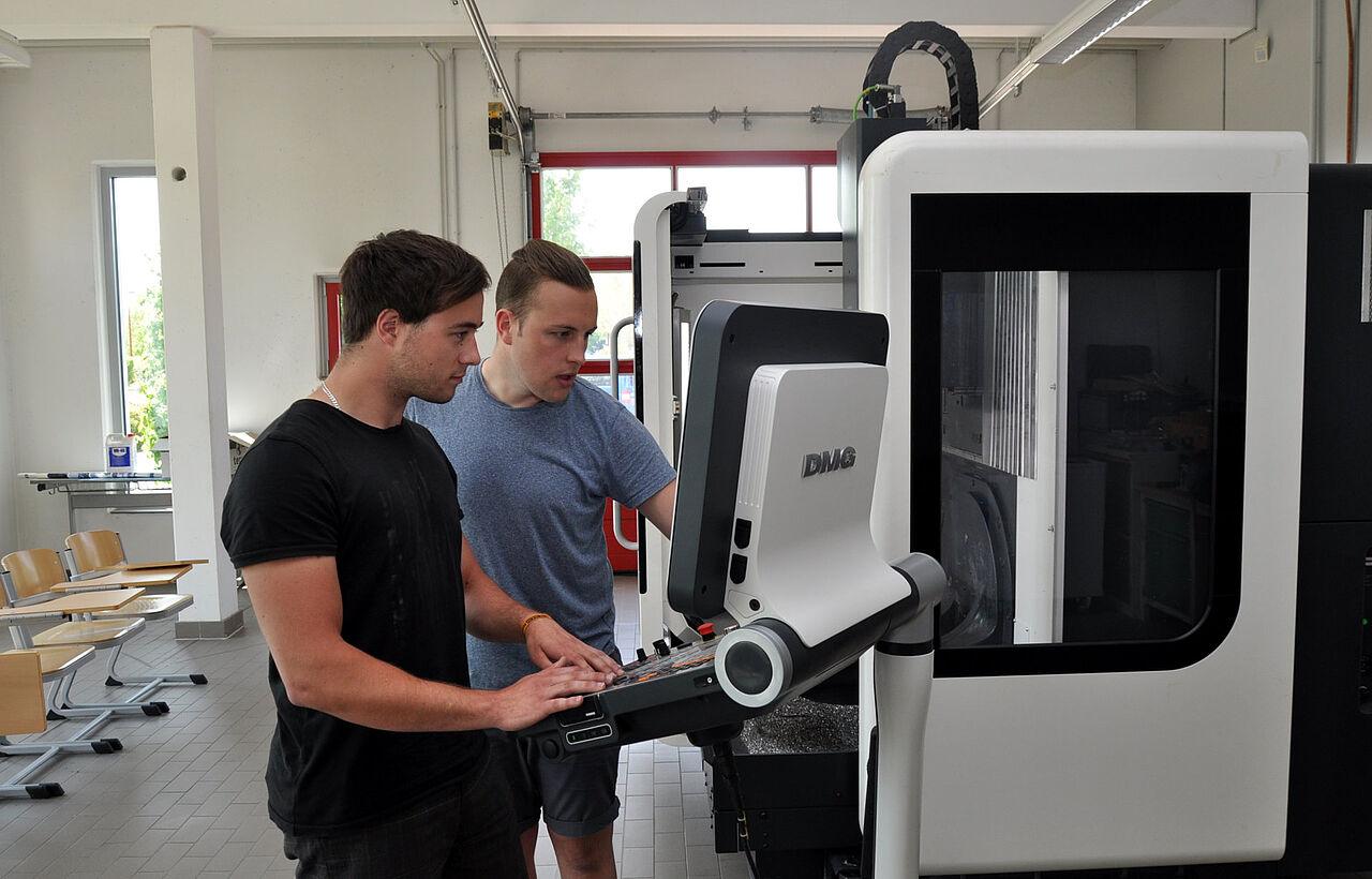 Zwei Auszubildende bedienen gemeinsam das Steuerpult einer CNC-Fräsmaschine in der Metallwerkstatt.