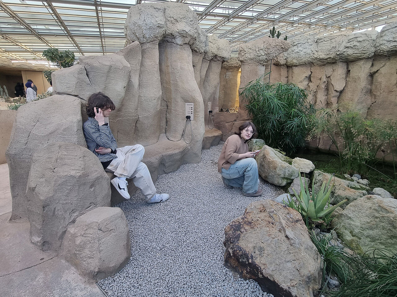 Zwei Schülerinnen und Schüler erkunden mit Kopfhörern und mobilen Geräten eine Felsen- und Pflanzenlandschaft im Grünen Klassenzimmer.