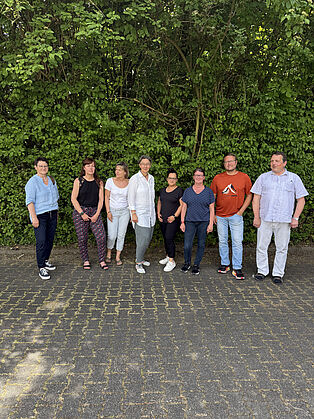 Acht Teilnehmende der Hospitation stehen für ein Gruppenfoto vor einer dicht bewachsenen grünen Hecke.