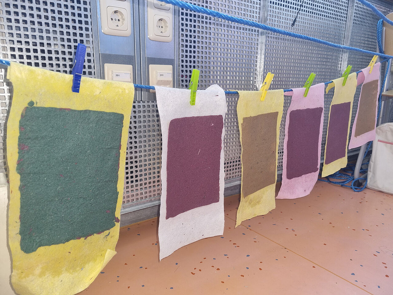 Mehrere farbige, selbst geschöpfte Papierbögen hängen mit Wäscheklammern an einer Leine und trocknen.
